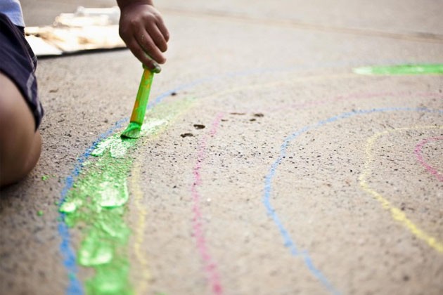 A child’s hand painting the first section of a rainbow shape sketched on the sidewalk.