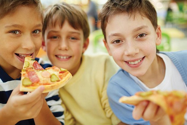 Three school-age boys eating pizza and smiling.